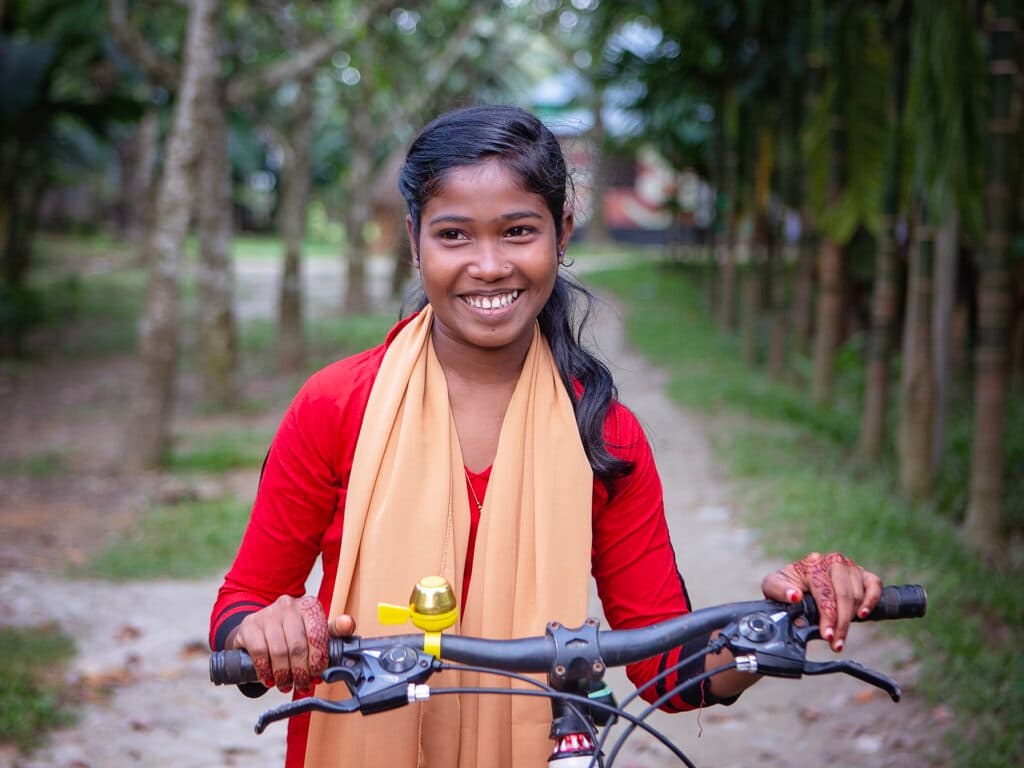 Foto: Diakonia En flicka med röd klänning och gul sjal står framför en cykel och ler.