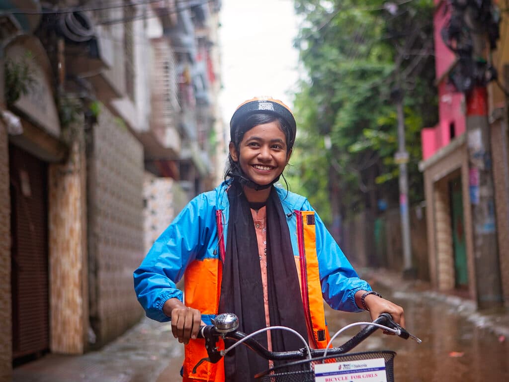 Foto: Diakonia En flicka i cykelhjälm och regnjacka står med en cykel framför sig på en regnig gata och ler.
