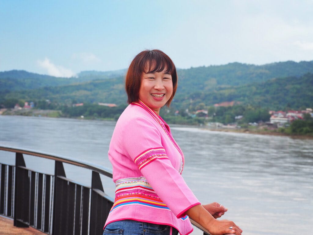 A woman in a pink blouse and brown hair looks into the camera, in the background there is a river.