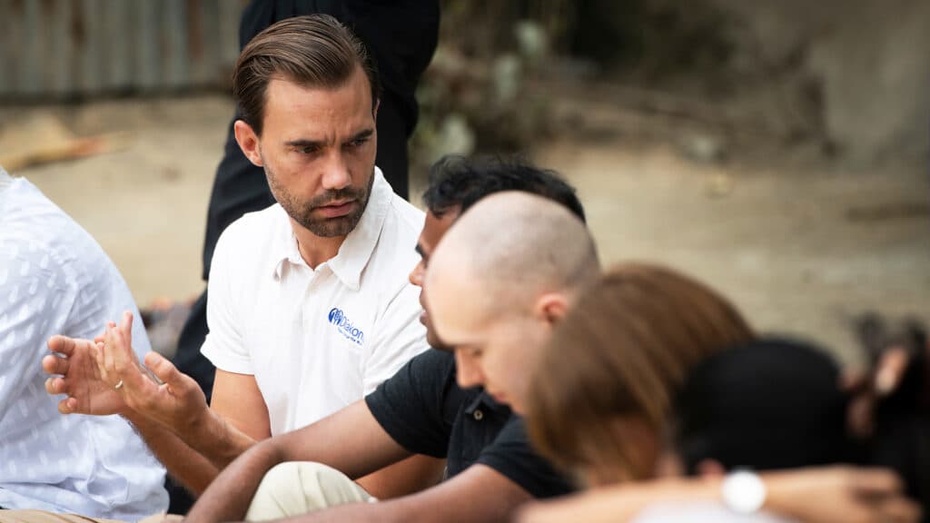 En man i vit t-shirt med Diakonias logo sitter i en grupp av människor och gestikulerar med händerna.