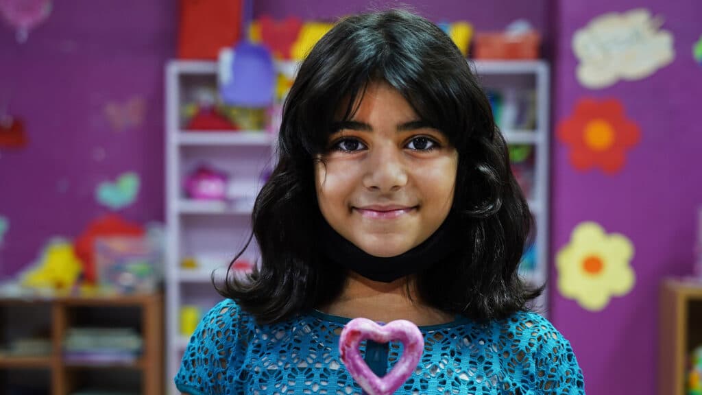 En leende flicka ser in i kameran. I bakgrunden syns lila väggar med målade blommor, leksaker och en bokhylla.