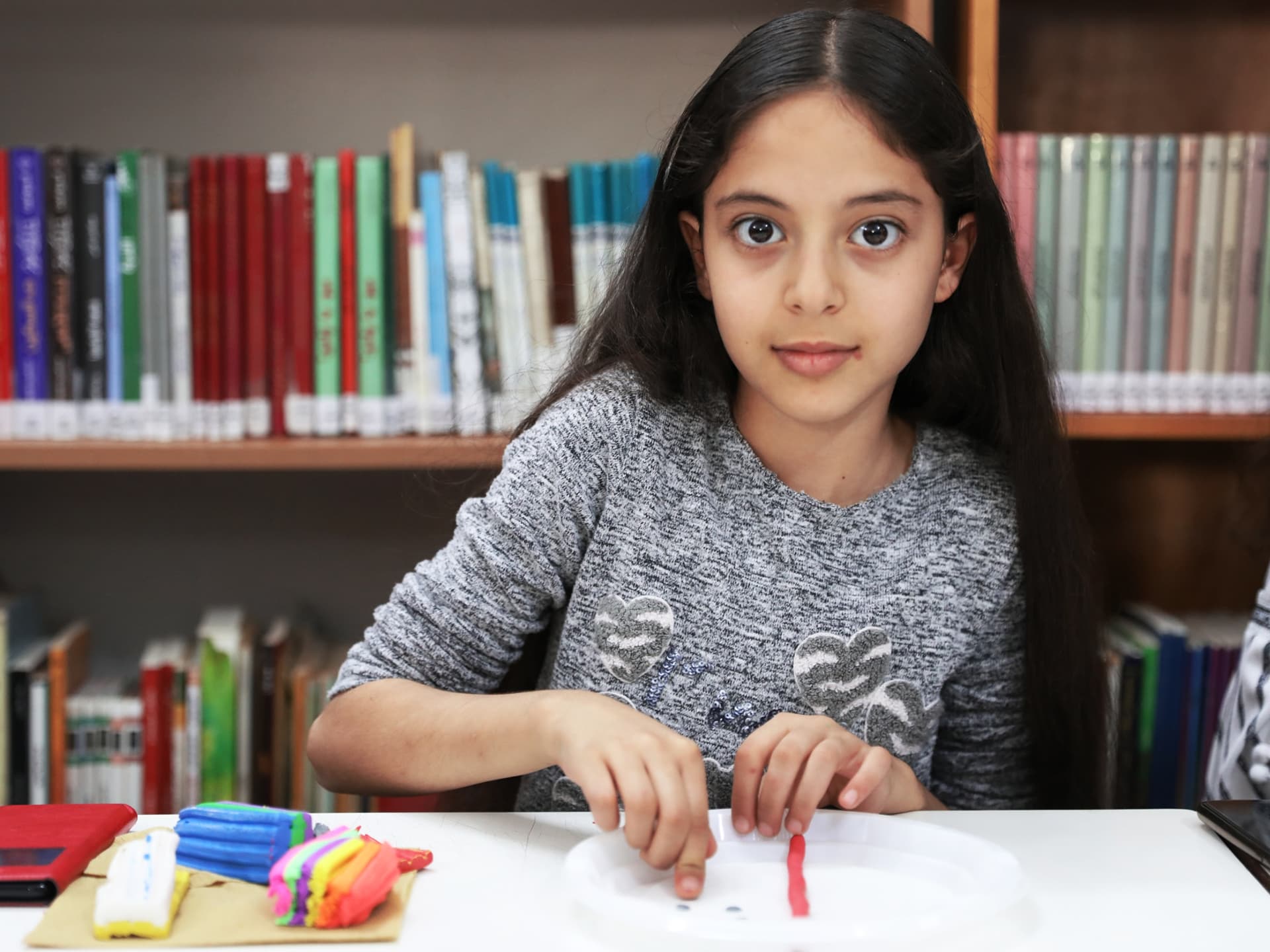Flicka sitter i ett bibliotek, framför sig har hon modellera i olika färg som hon jobbar med.