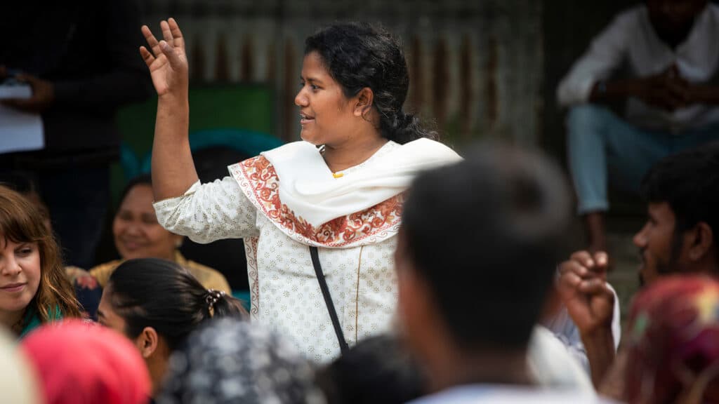 Kvinna som står upp i en grupp av människor. Hon pratar och gestikulerar med handen.