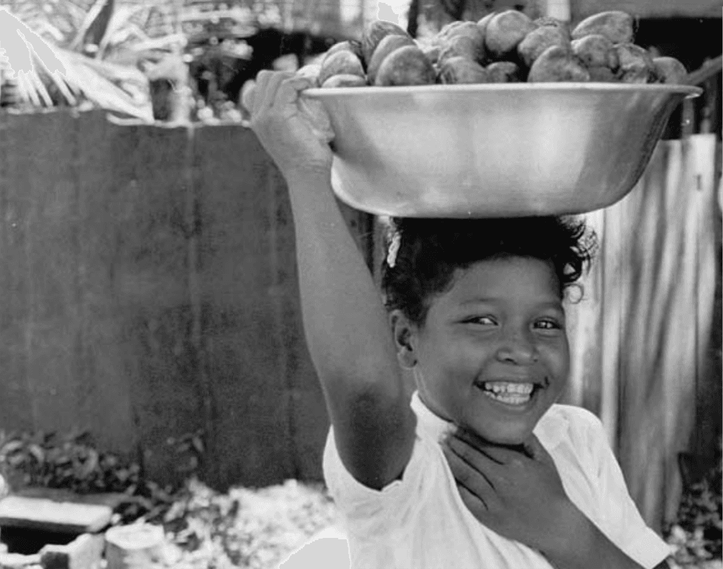 Ett svartvitt foto av ett leende barn som bär ett kärl på huvudet.