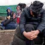 Två barn sitter på marken med framför ett stängsel där ett täcke hänger.