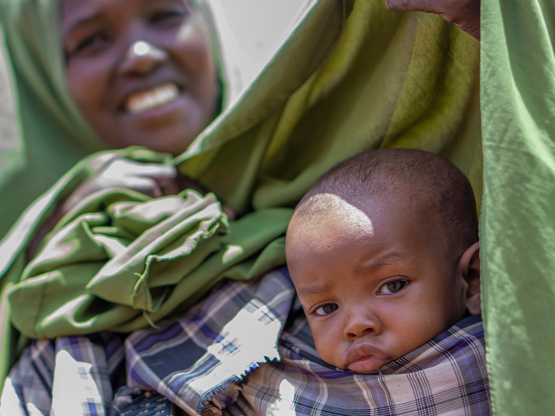 Ett litet barn i framgrunden ser in i kameran. Bakom barnet syns en leende kvinna och ett grönt tygstycke.