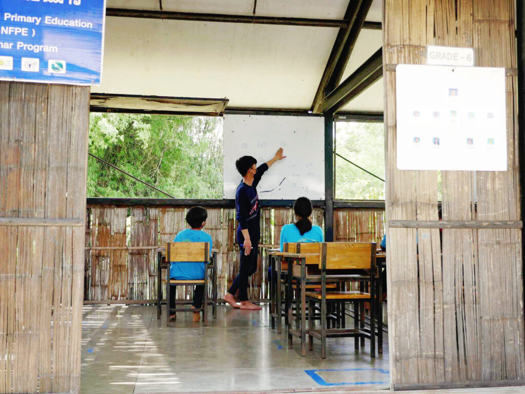 En grupp med barn sitter i skolbänkar med ryggarna vända mot kameran. Vid en tavla står en man och pekar.