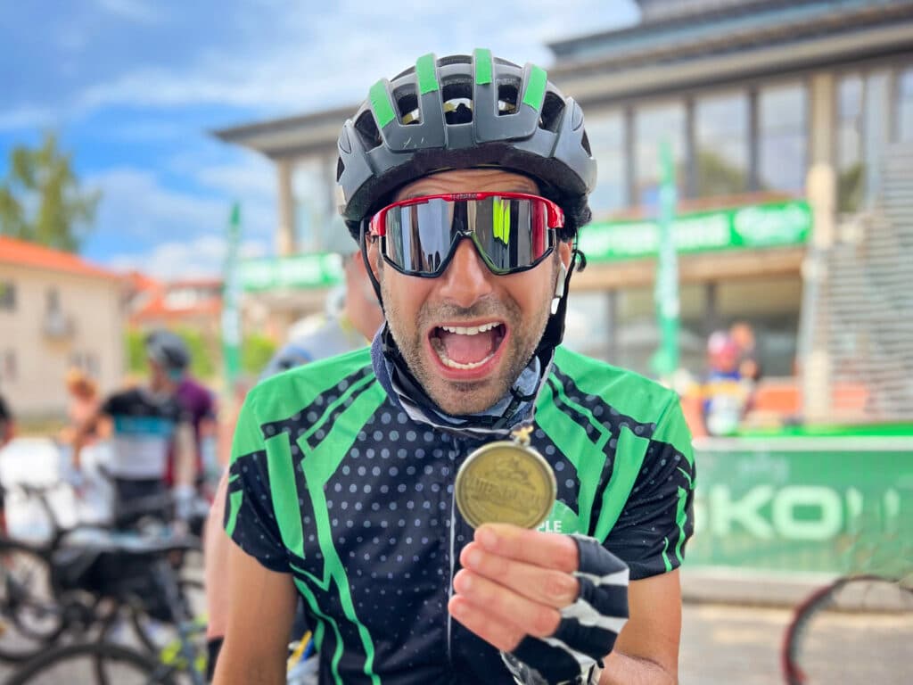En bild av Stephan Wilson i solglasögon, cykelhjälm och med cykelhandskar som håller upp en medalj framför kameran.
