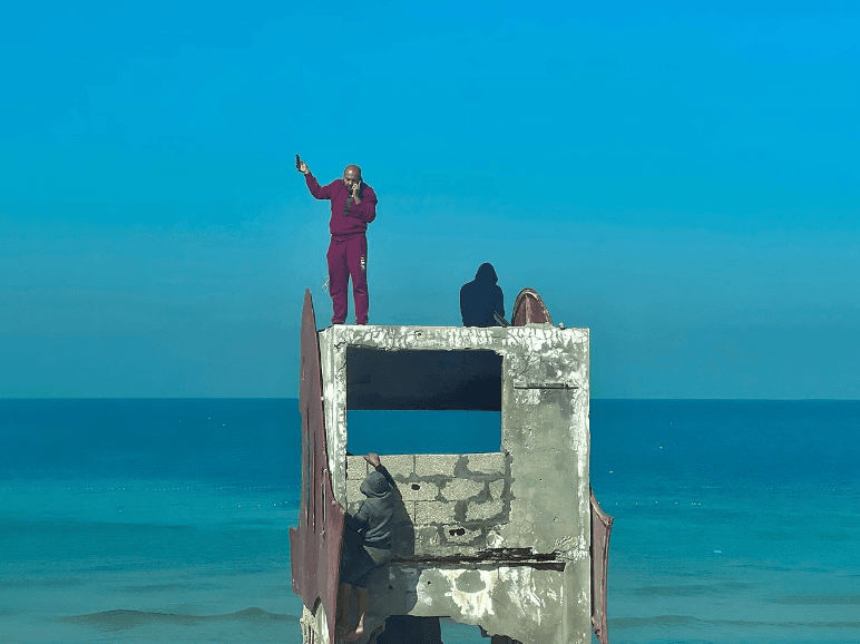 En man står högst upp på ett trasigt hustak med en mobiltelefon i handen, som han sträcker upp i luften.