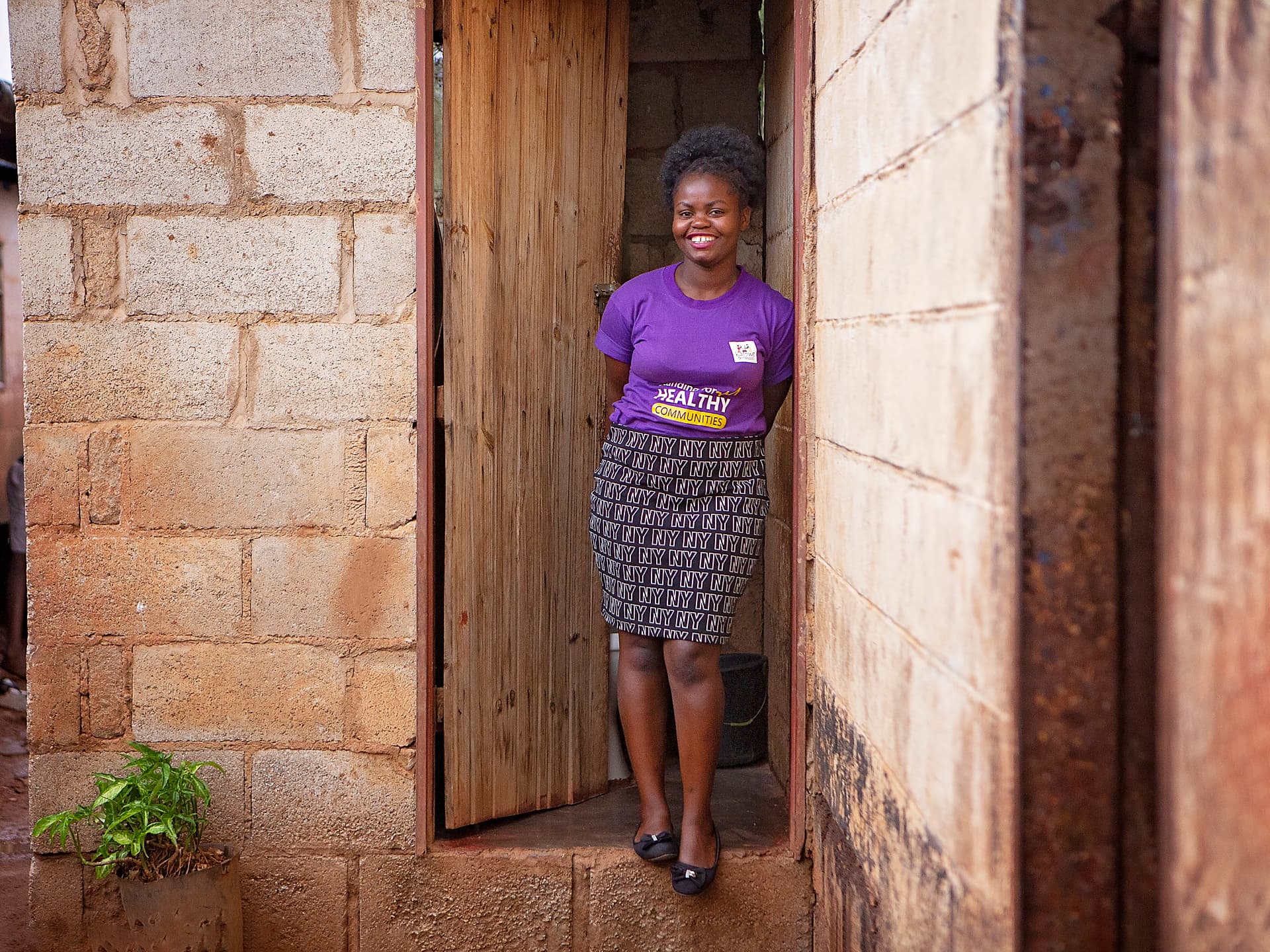 En kvinna i lila t-shirt och kjol står i en dörröppning i ett hus av tegelsten och lera.