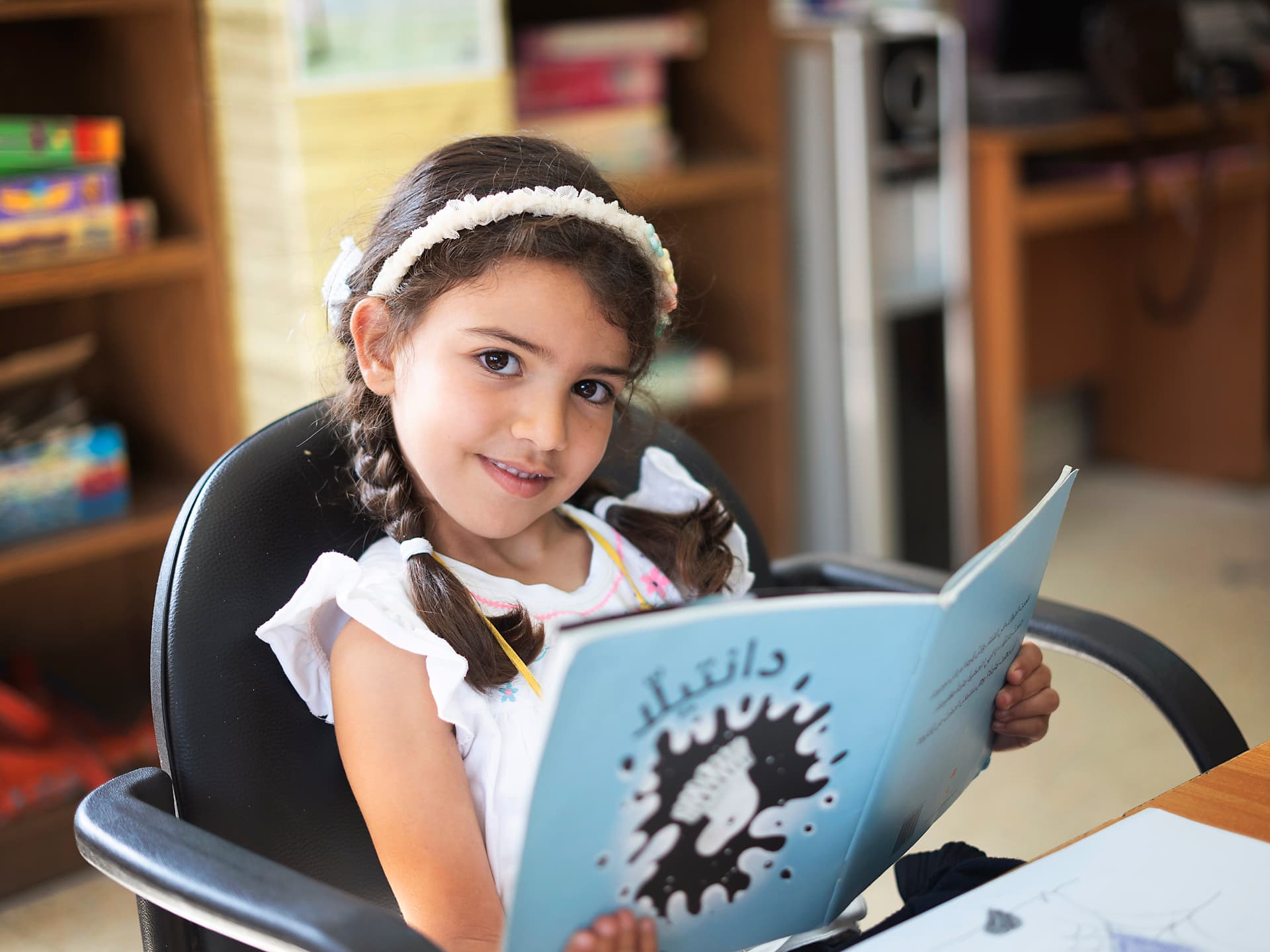 En flicka med flätor sitter i en stol med en barnbok i handen.