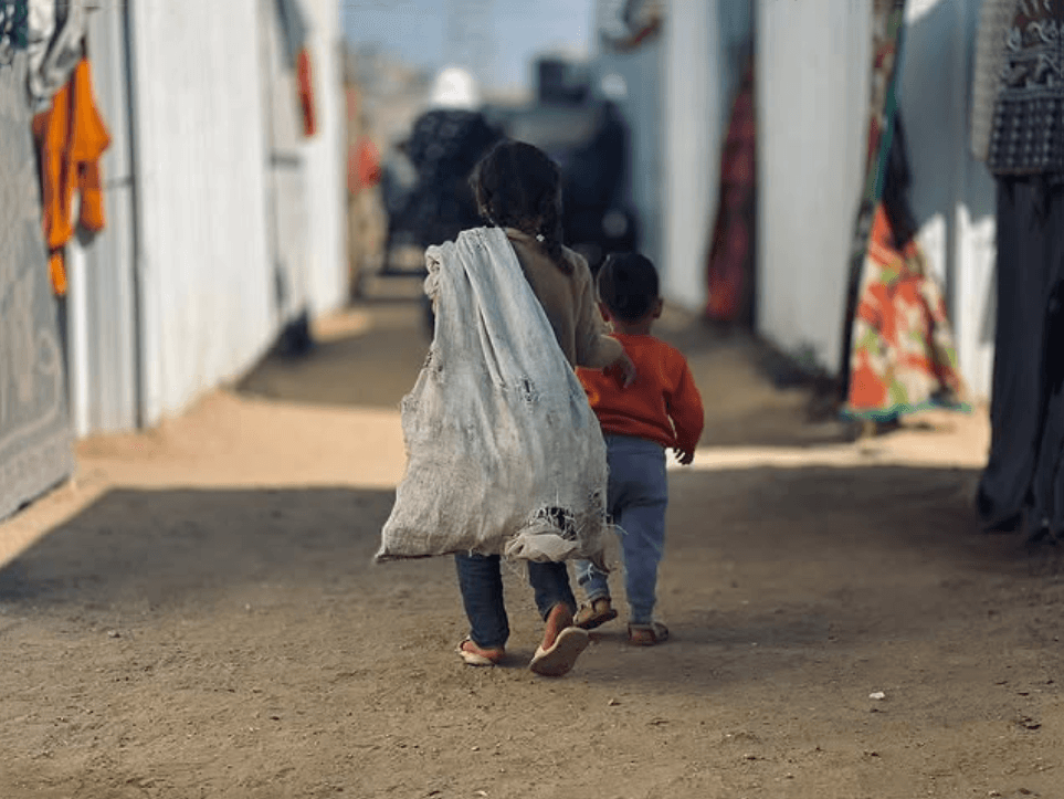 Två barn promenerar på en jordväg med ryggen mot kameran. De bär på en stor vit säck.