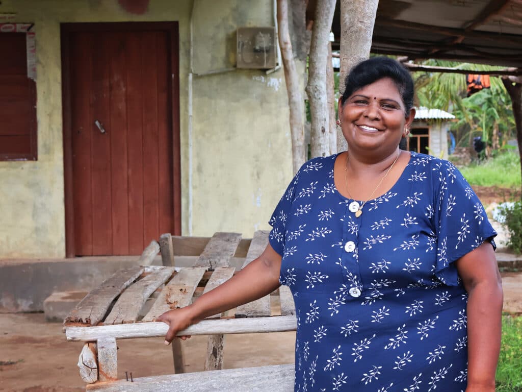 A darkhaired woman in a blue blouse stands in front of a house, smiling.