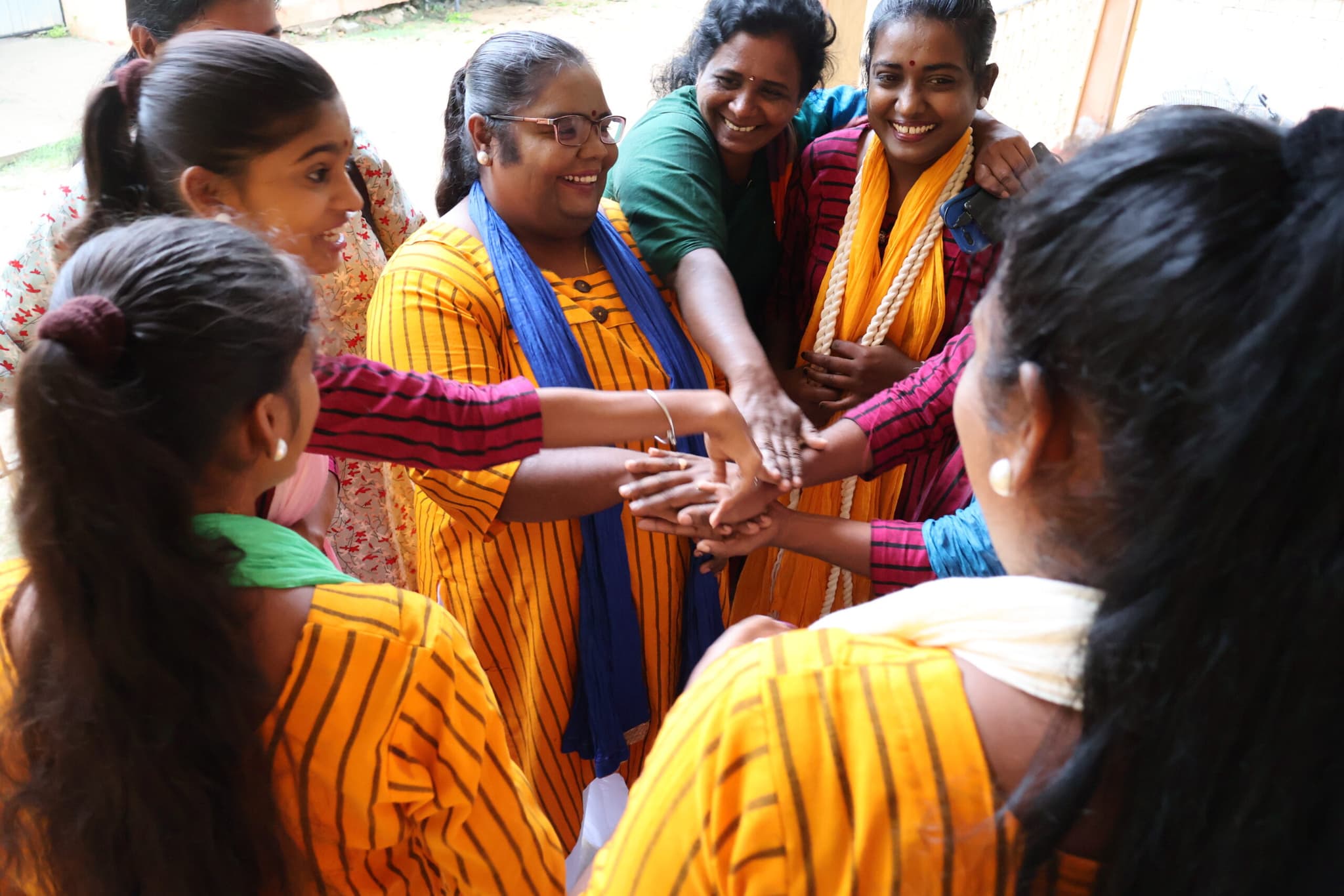 Sex glada kvinnor i färgglada kläder som står i en ring och lyfter in sina händer mot mitten av ringen.