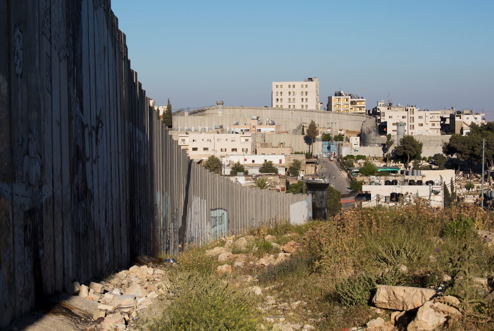A high concrete wall runs through a valley, with houses next to the wall.