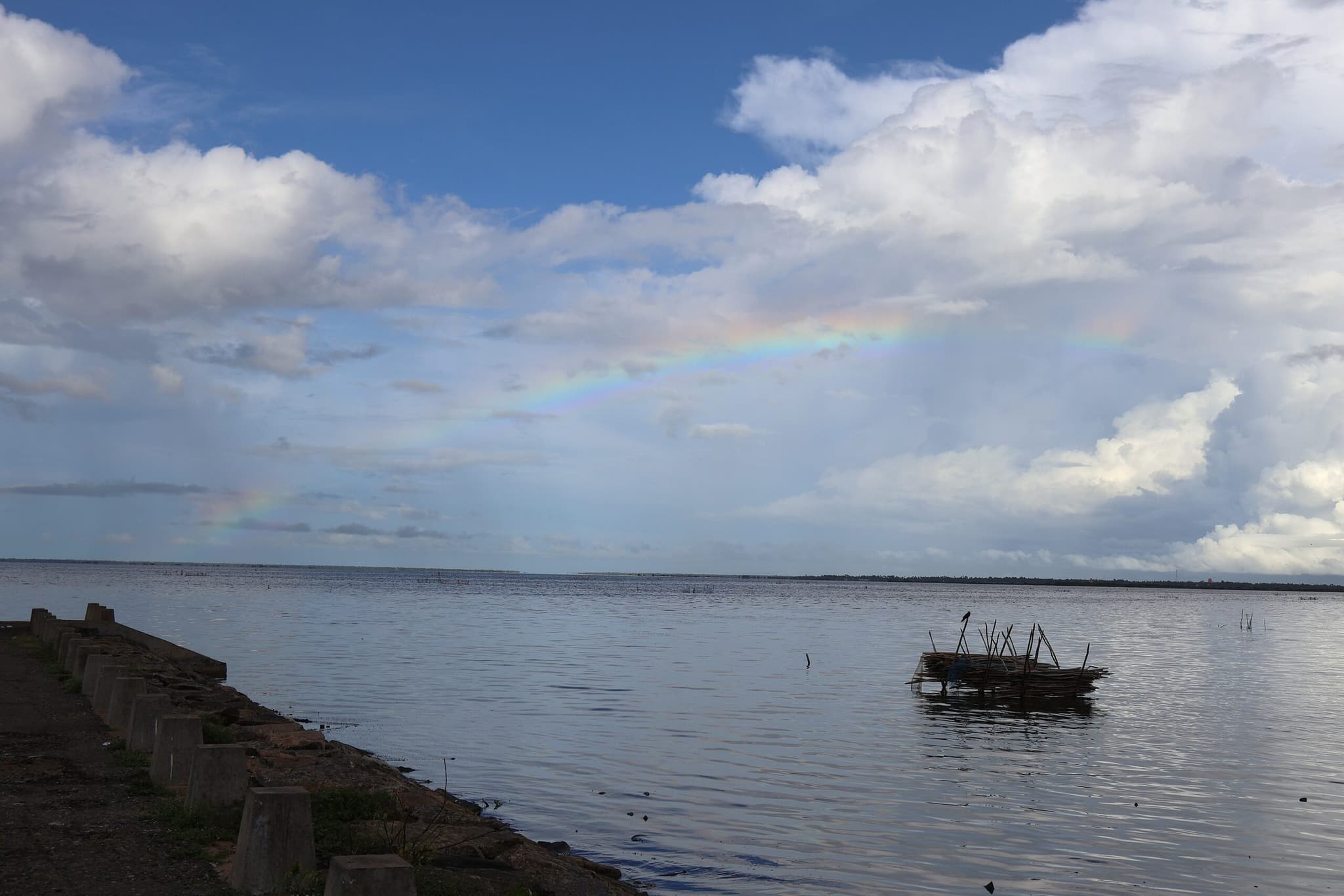 Wide view of a lagoon under a cloudy blue sky, with still water and a small wooden structure floating near the shore.