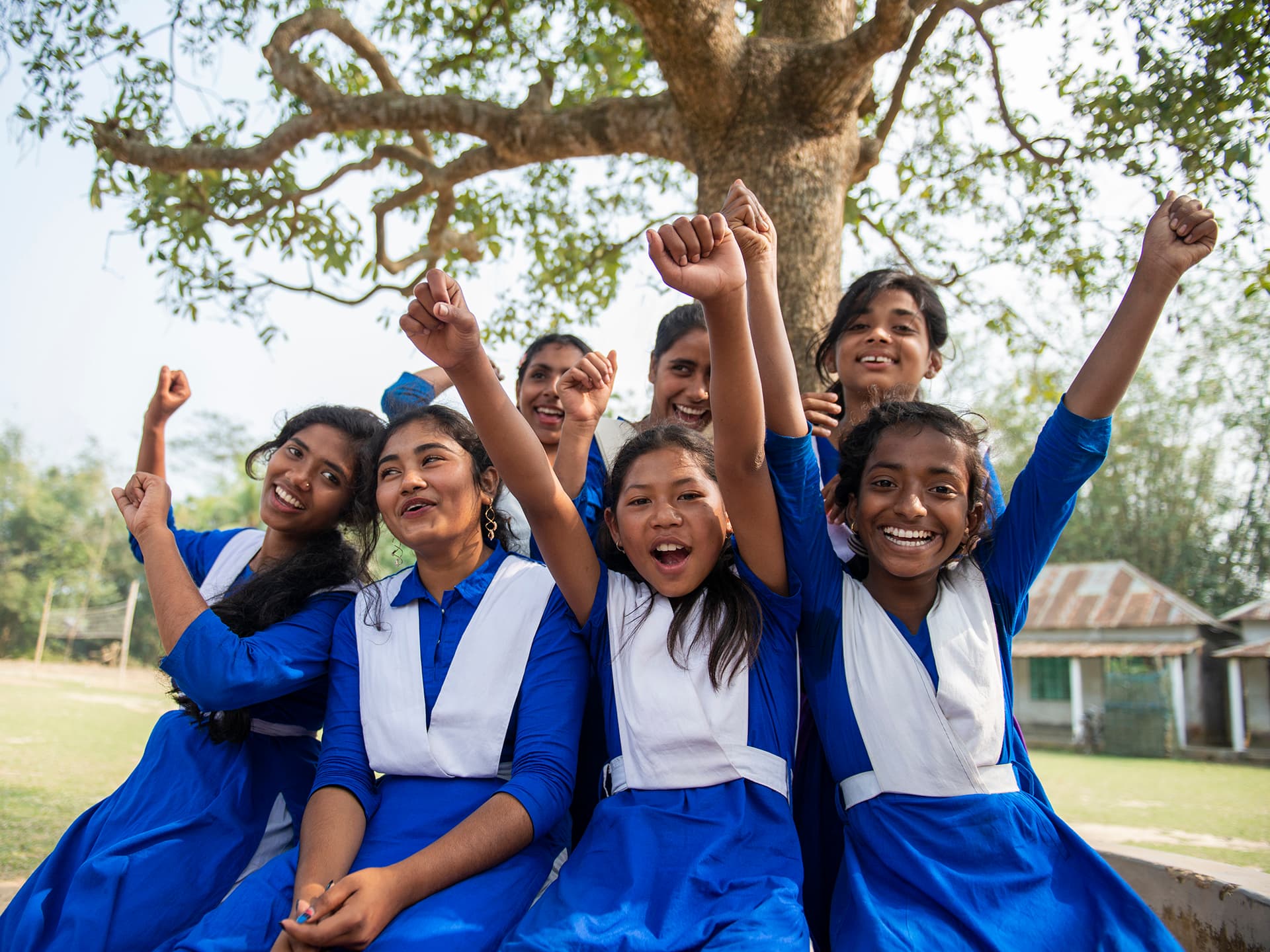 Foto: Gustav Hugosson En grupp av leende och skrattande flickor sitter intill ett träd. De är klädda i likadana blåa skoluniformer och sträcker upp händerna i luften.