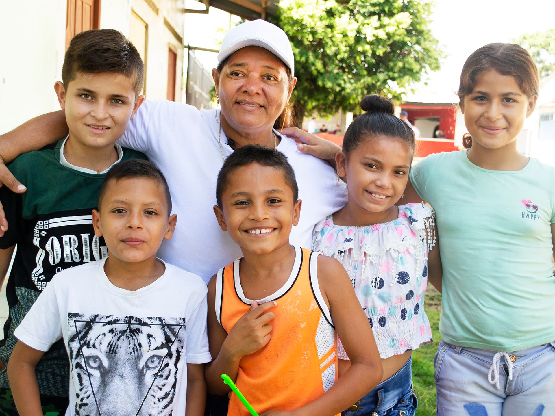 Foto: Diakonia En kvinna i keps står med armen om fem barn. I bakgrunden syns ett träd och en skolbyggnad.