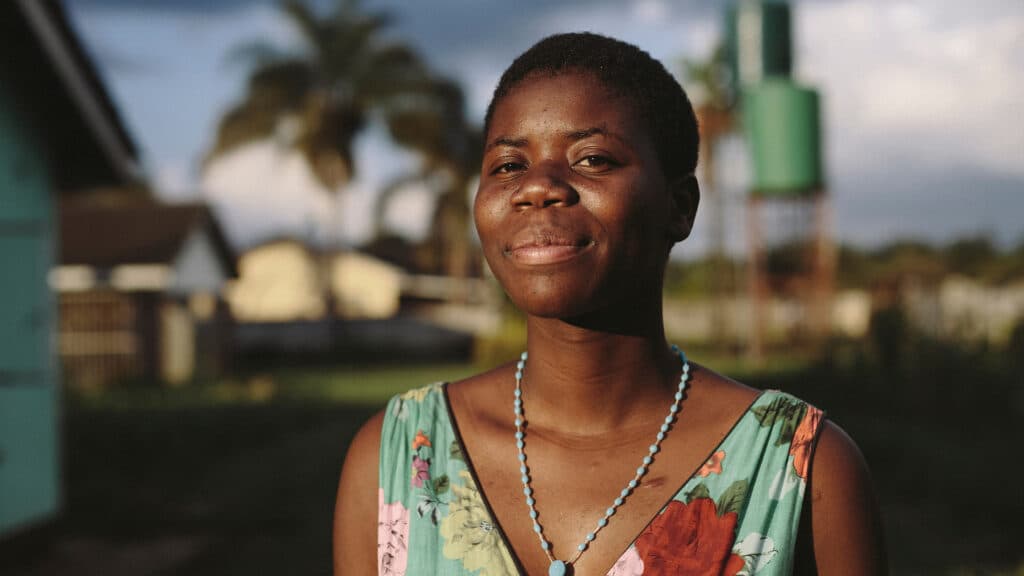 Kvinna i Zimbabwe tittar in i kameran. I bakgrunden syns låga byggnader och fält av gräs.