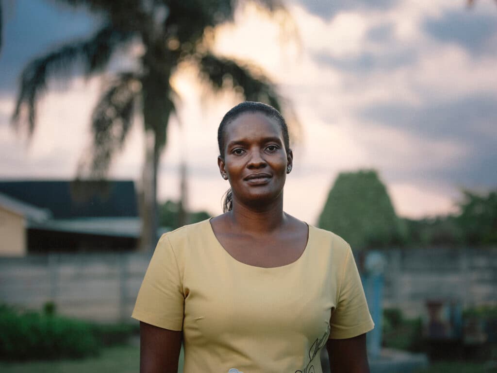 Foto: Anna Hugosson Bild av en kvinna med gul tröja som ser in i kameran. Bakom henne syns låga byggnader och gröna träd.