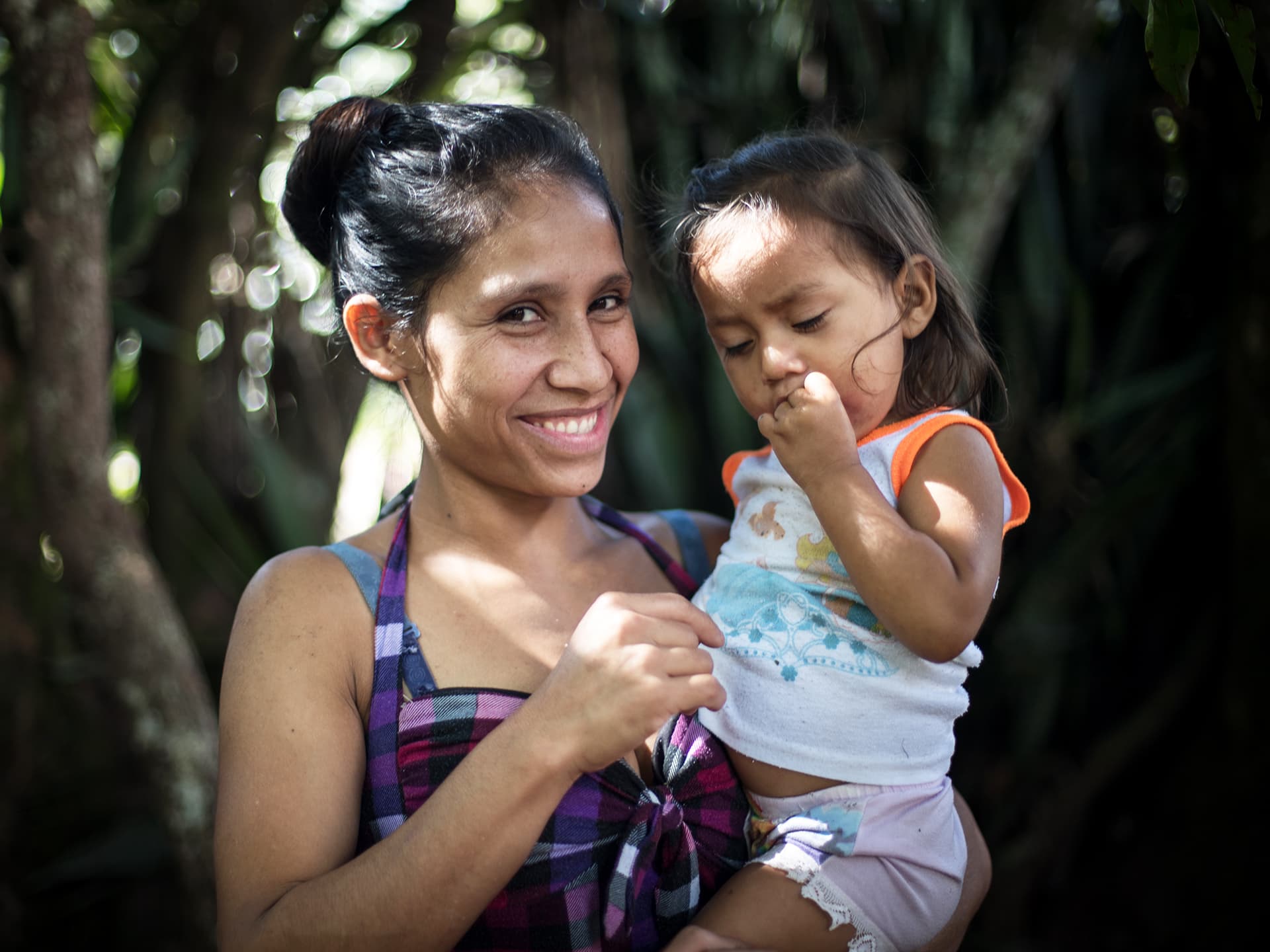 Foto: Stephen Hawkey En leende mamma står med ett litet barn i famnen framför träd och buskage.