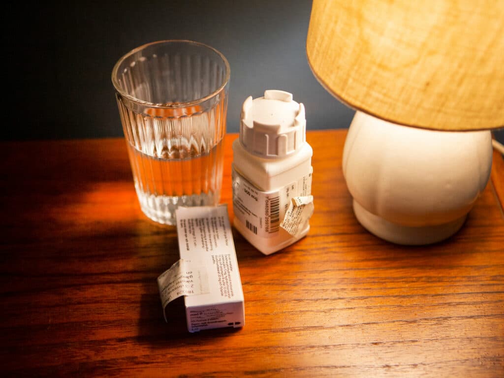 Ett nattduksbord med ett glas, medicinpaket och en lampa.