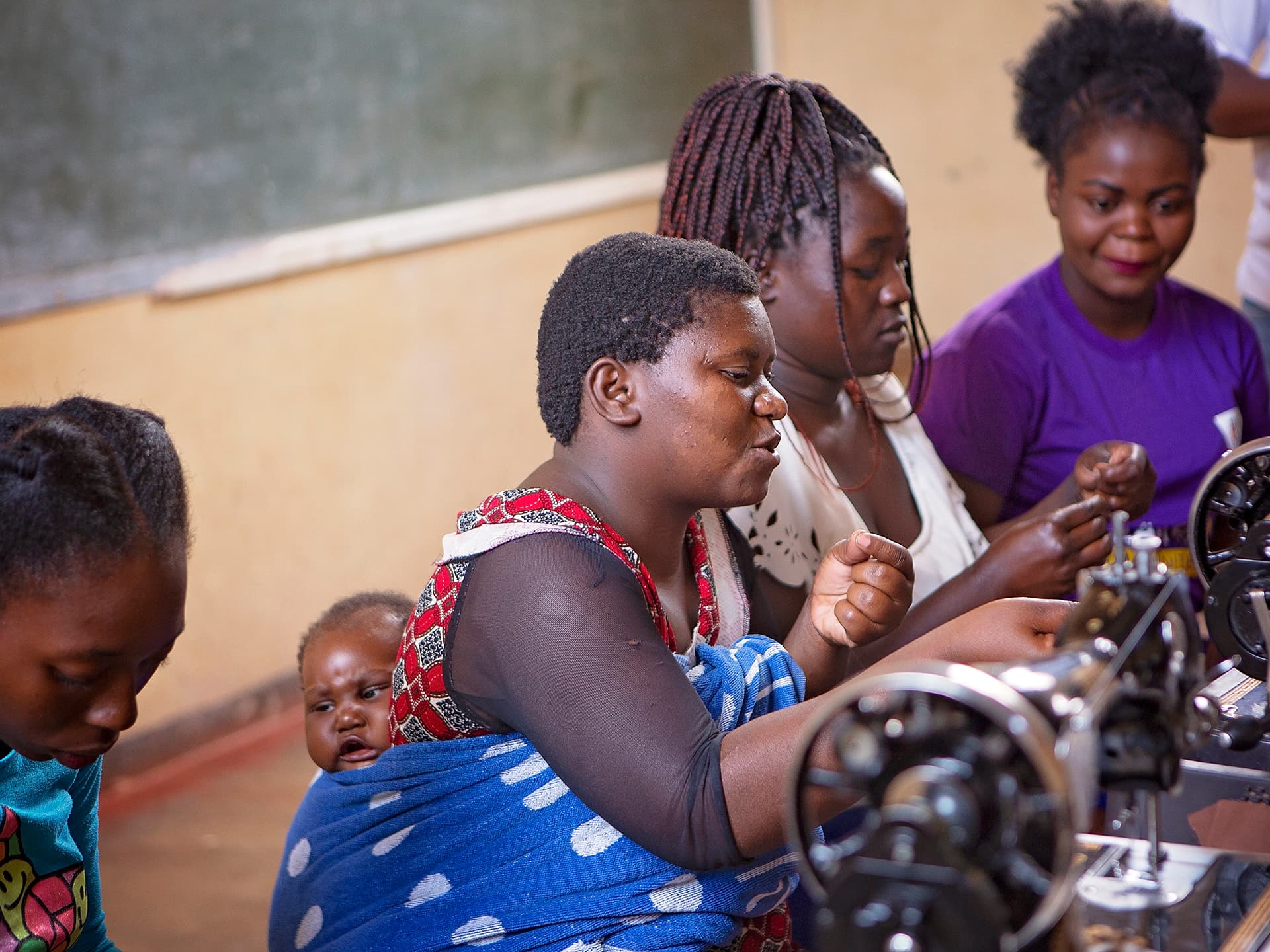 Foto: Diakonia Fyra unga kvinnor sitter vid symaskiner. En av kvinnorna har en bebis i en sjal på ryggen.
