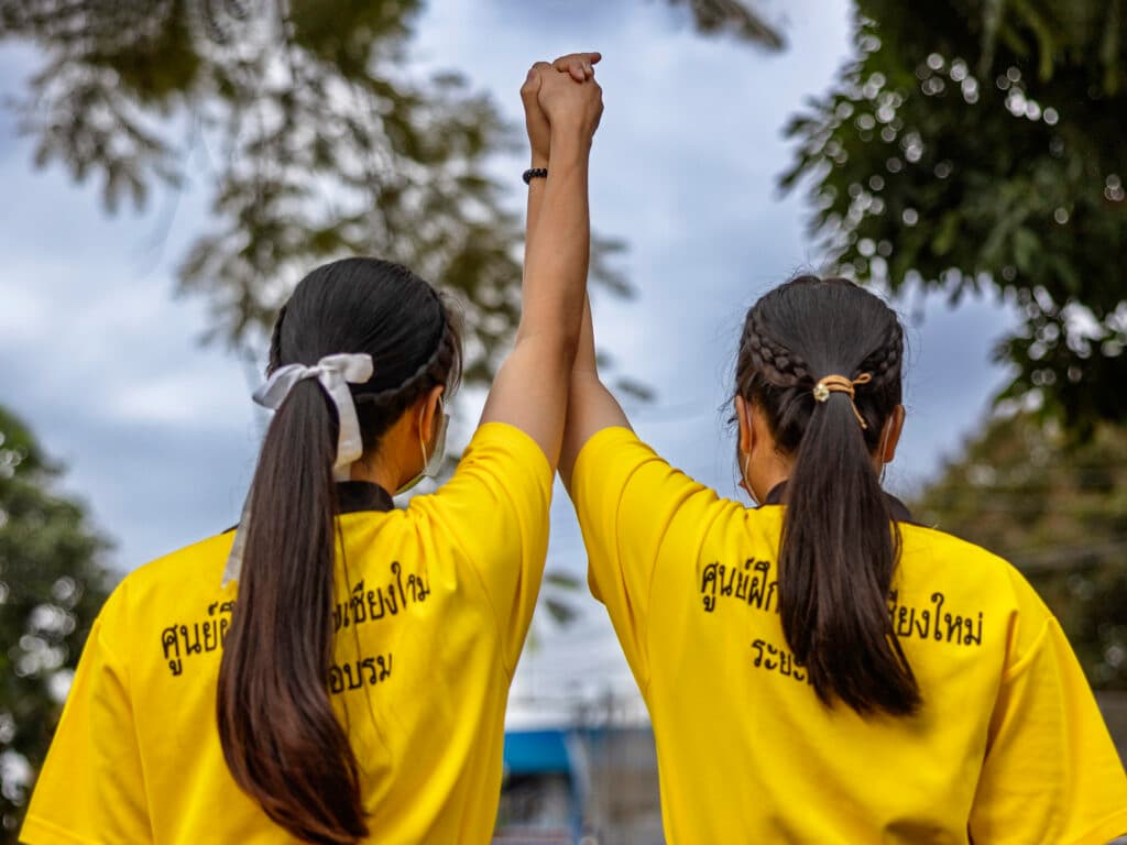 Två unga kvinnor med likadana gula tröjor står med ryggarna mot kameran och håller sina händer upp mot himlen.