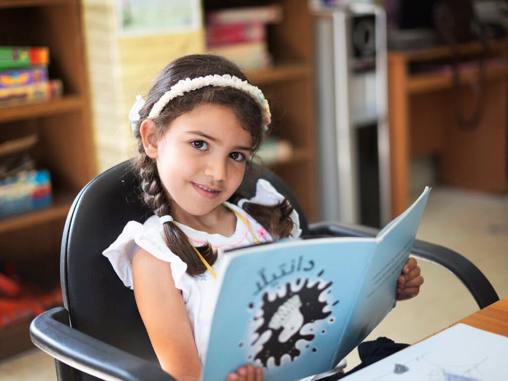 Foto: Ahmad Daghlas En flicka med flätor sitter i en stol med en barnbok i handen.