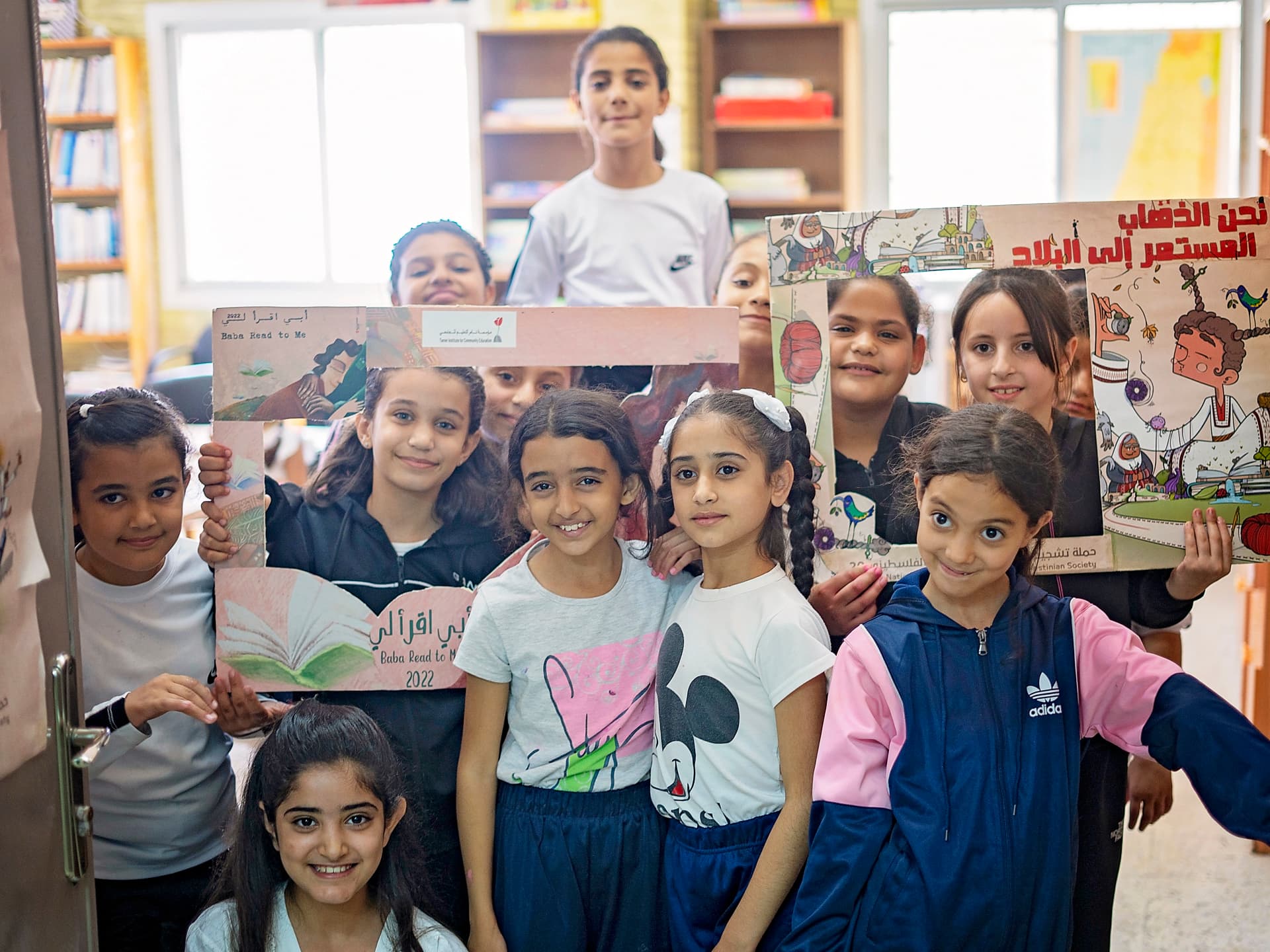 Foto: Ahmad Daghlas En grupp barn i ett bibliotek håller upp teckningar mot kameran och ler.
