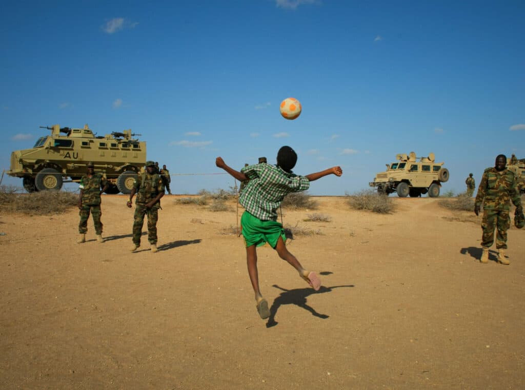 A boy playing with a ball, with soldiers and military vehicles in the background. A boy playing with a ball, with soldiers and military vehicles in the background. Photo: AU-UN IST PHOTO / Stuart Price, CC BY-SA 2.0