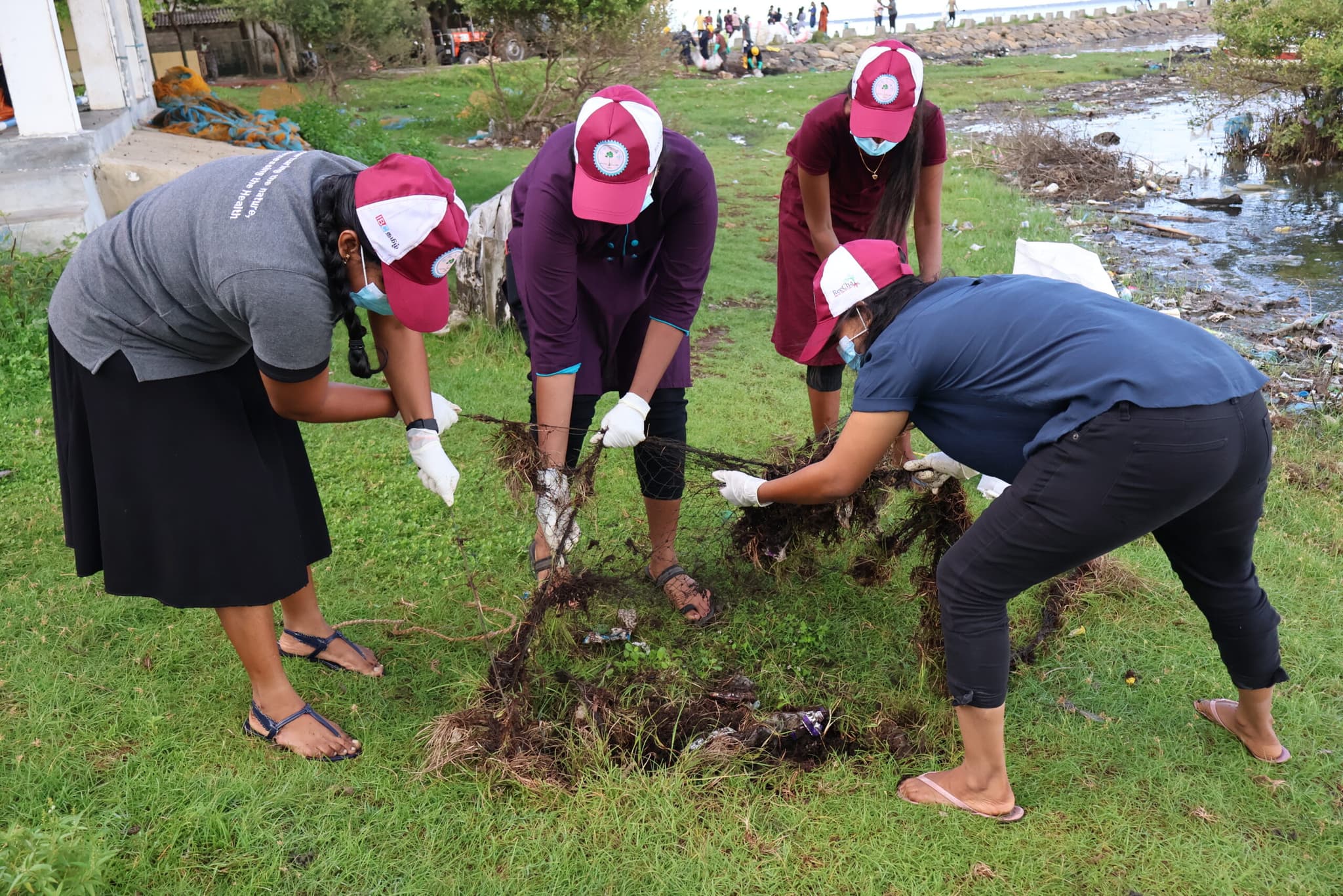 Women volunteers work together to clean and remove debris from grass near a shoreline during a community clean-up. Four women wearing caps and gloves remove debris from grass near a shoreline during a community clean-up.