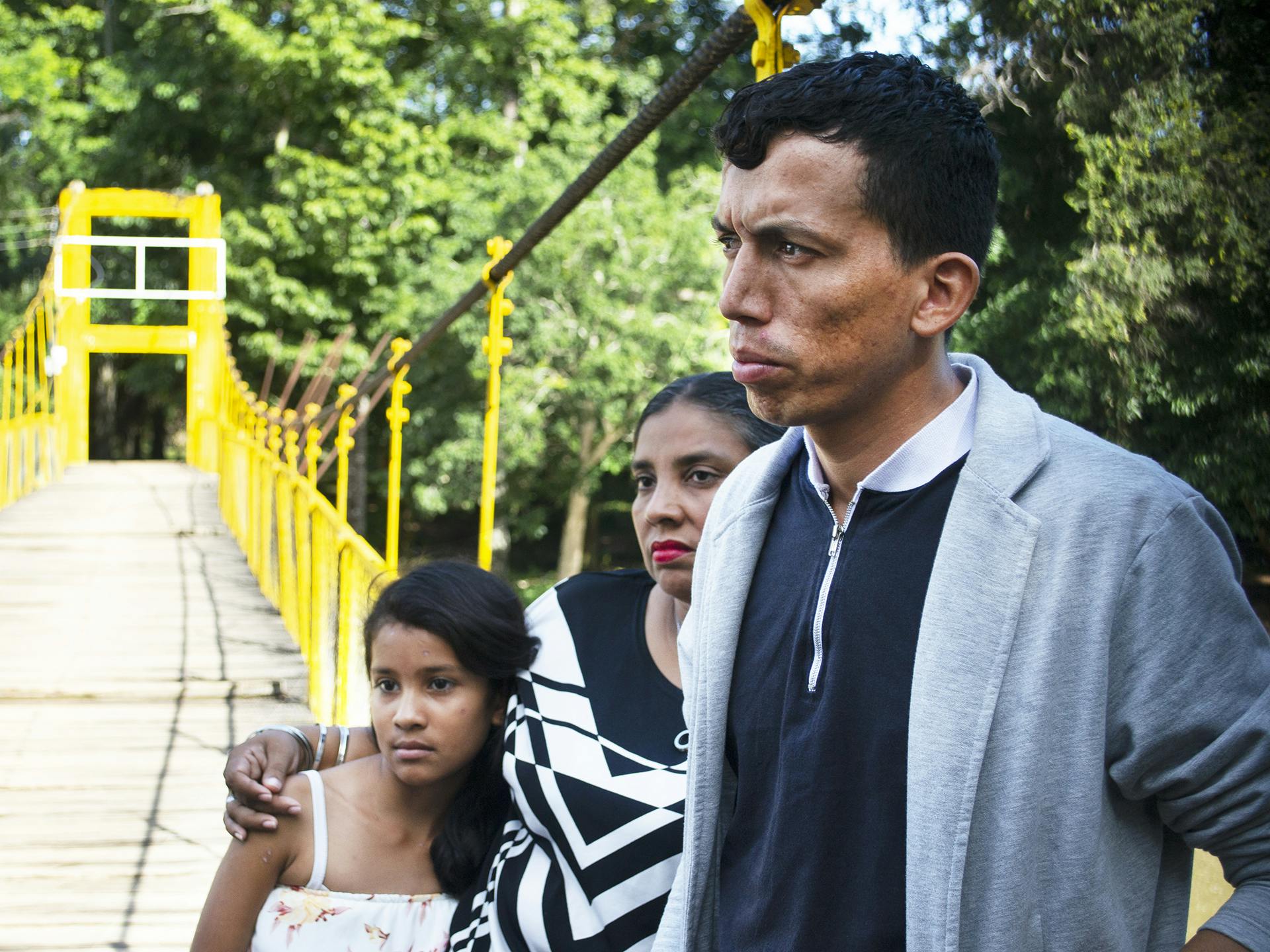 Man står med barn och kvinna vid bro, skog syns i bakgrunden