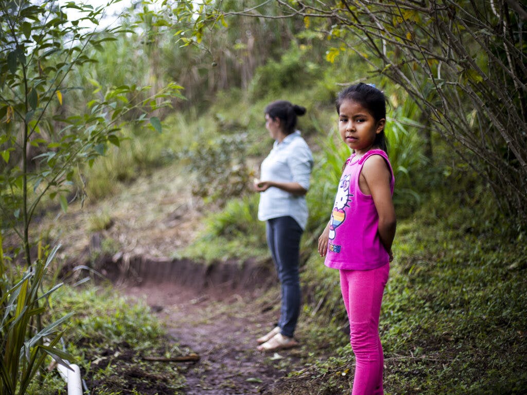 Ett barn står längst fram i bilden och tittar mot kameran, på en skogsstig. Bakom barnet står en kvinna.