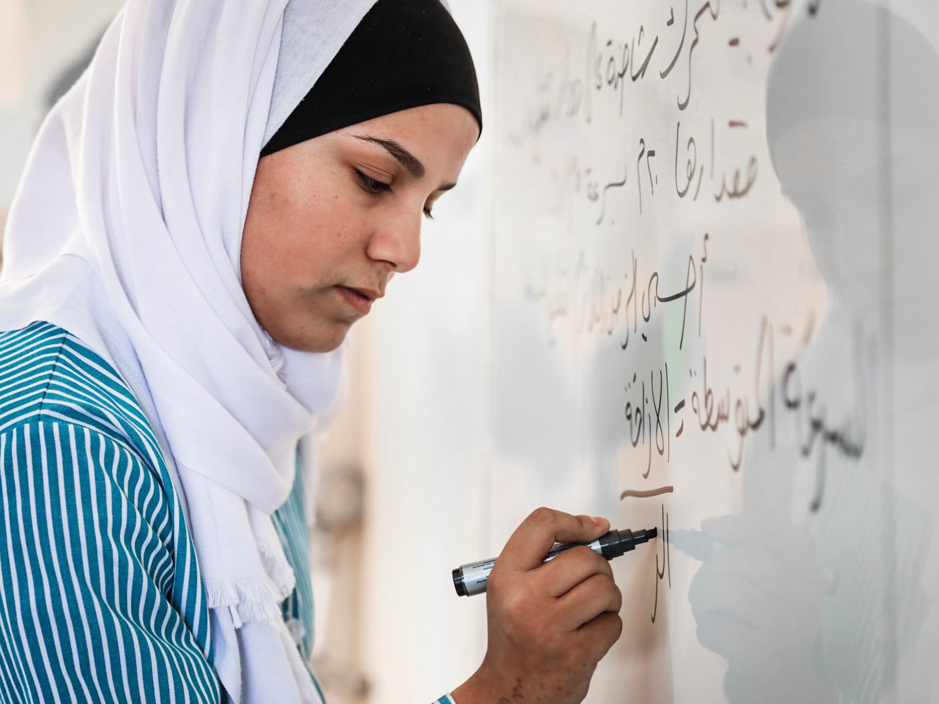 En kvinna i vit hijab skriver med svart penna på en whiteboard.