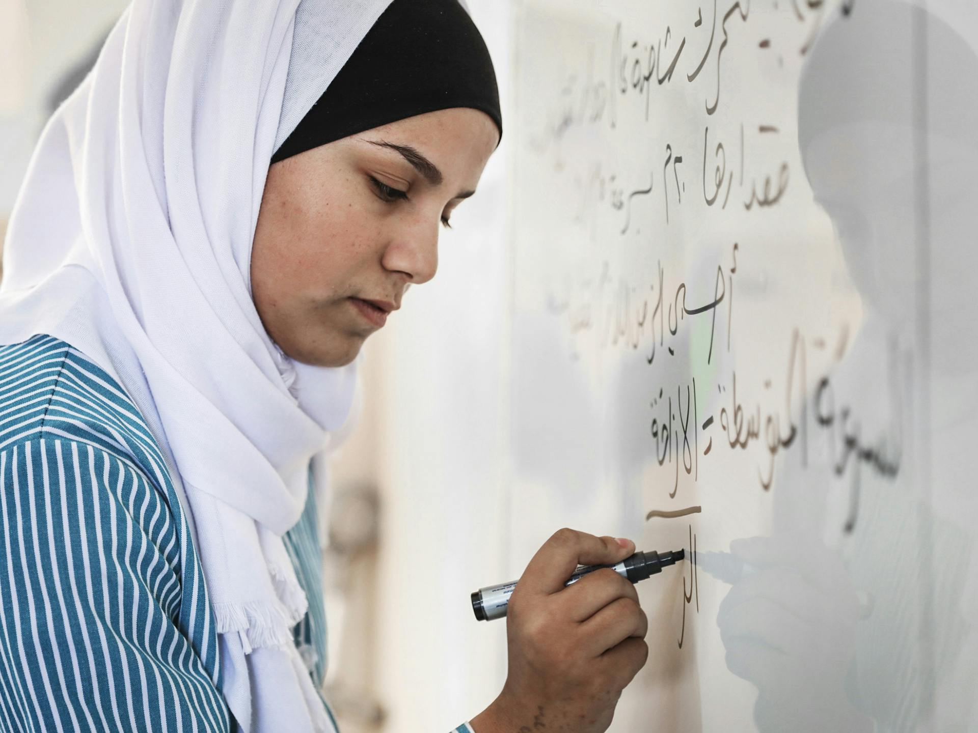 En kvinna i vit hijab skriver med svart penna på en whiteboard.