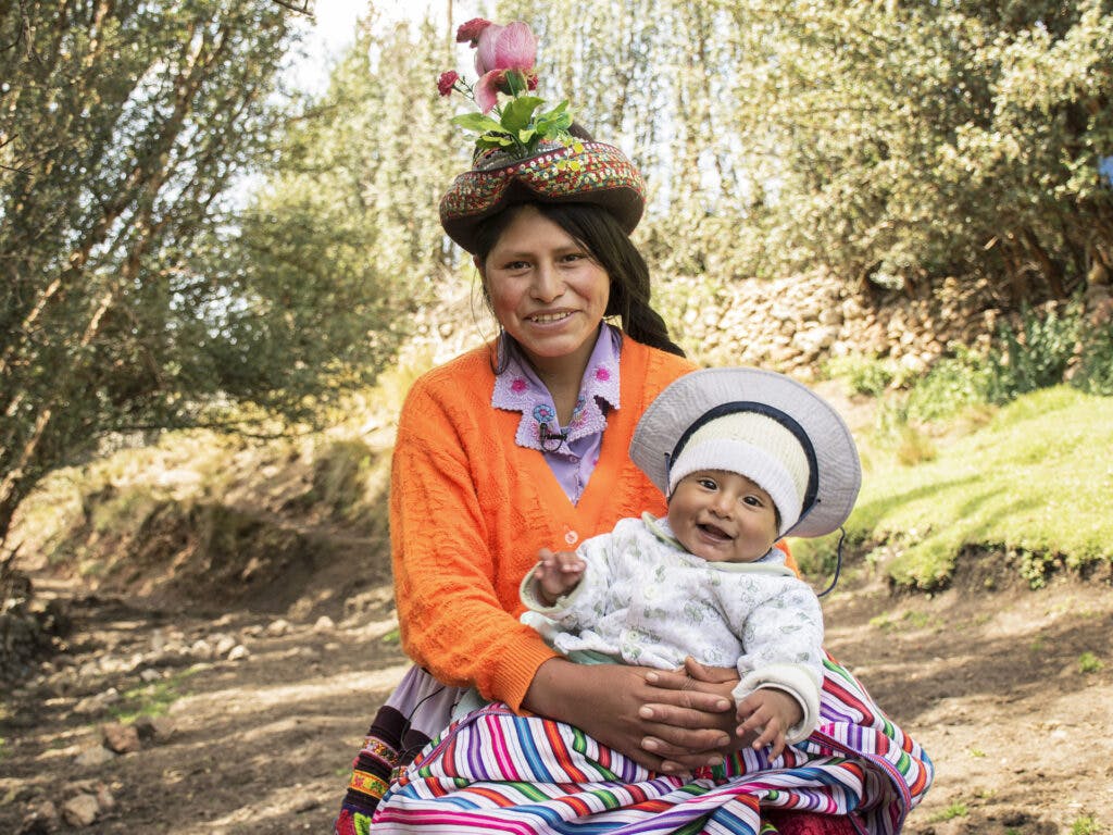 En leende kvinna sitter i ett grönområde med en bebis i famnen.