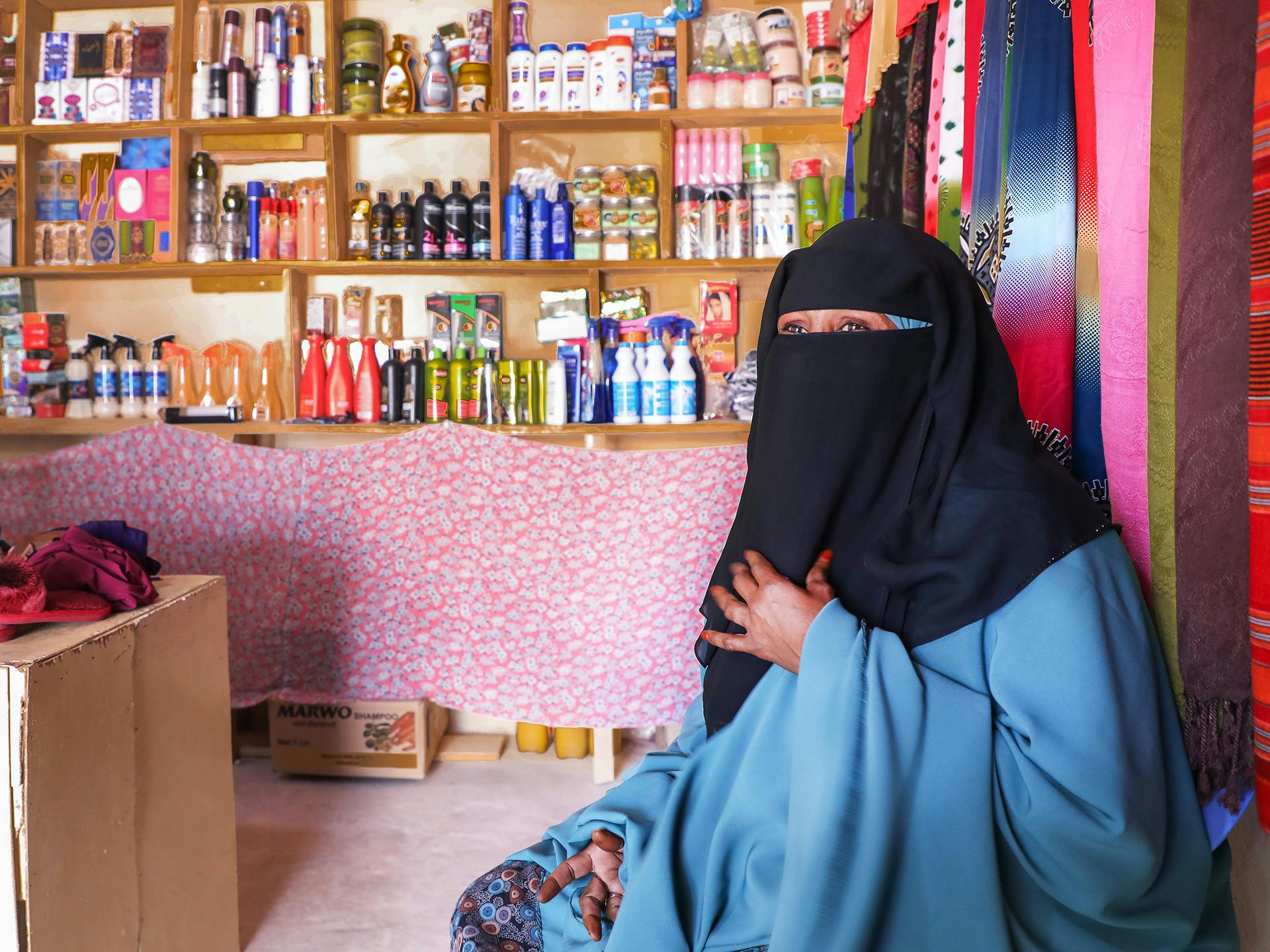 En kvinna i svart niqab och blå hiijab sitter på golvet i en butik. I bakgrunden syns hyllor med livsmedelsprodukter och tyger i olika färger och mönster.