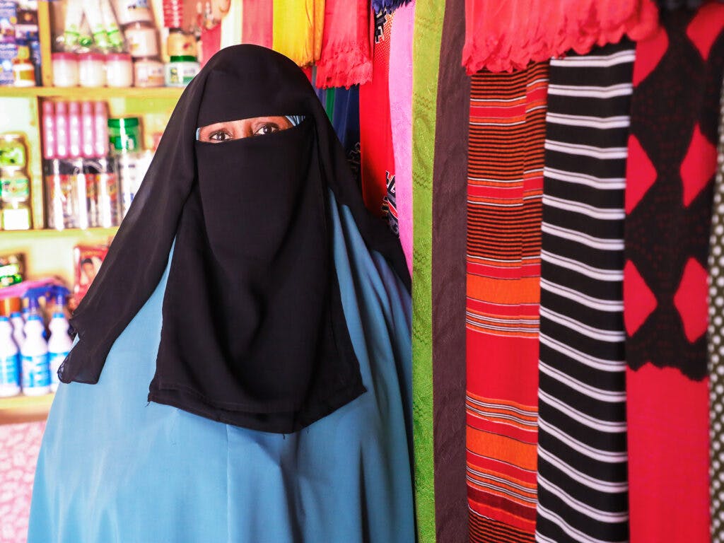 Porträtt av kvinna som bär svart niqab med en blå hijab under. Hon står lutad mot en vägg där det hänger tyger i olika mönster och färger.