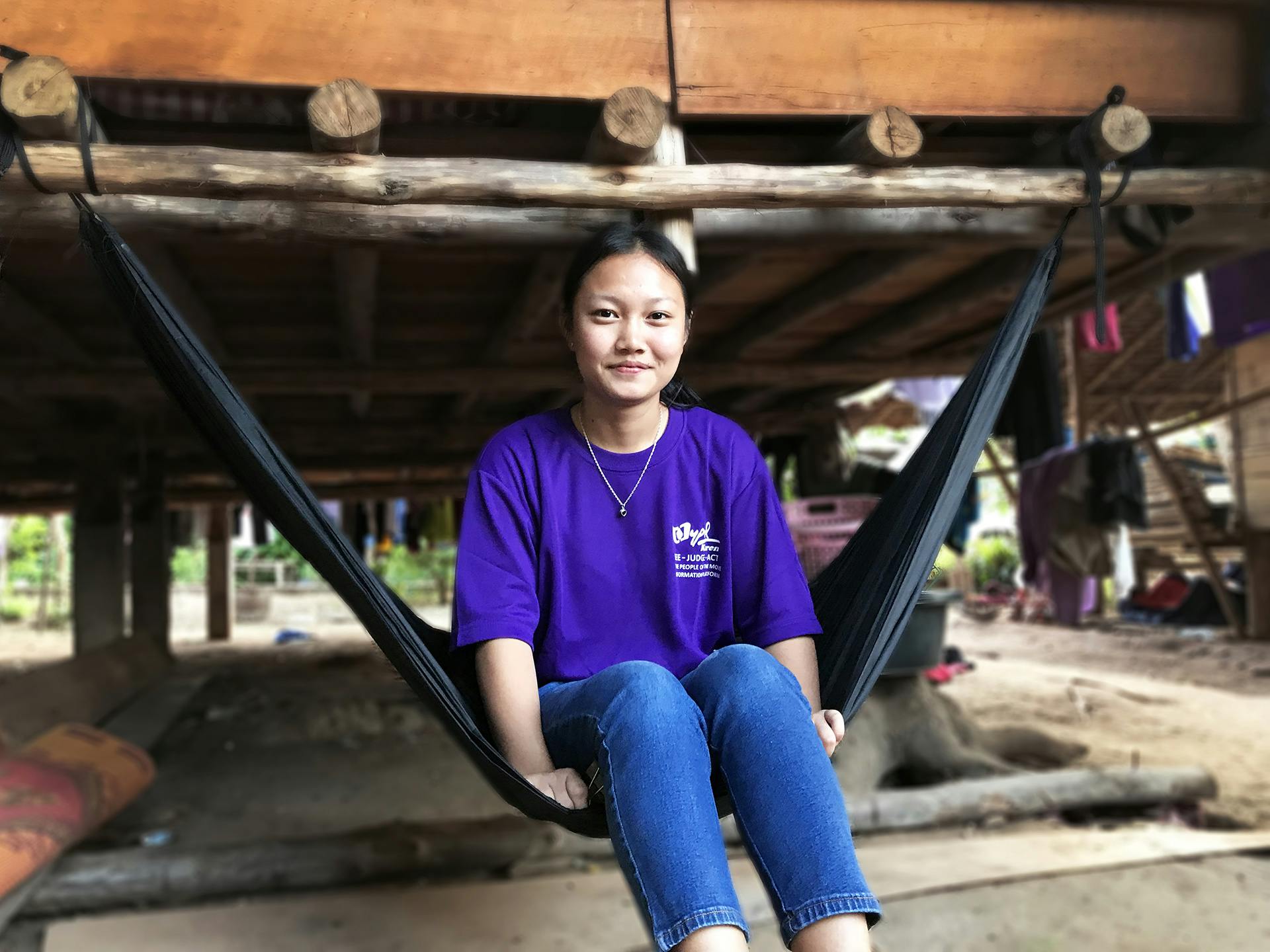 Ung kvinna i lila t-shirt och jeans sitter i en hängmatta. Bakom henne syns en grunden av en byggnad.