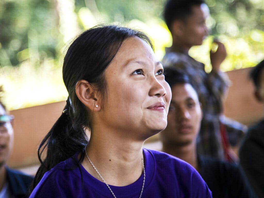 Närbild av ung leende kvinna i lila t-shirt. I bakgrunden syns fler människor.