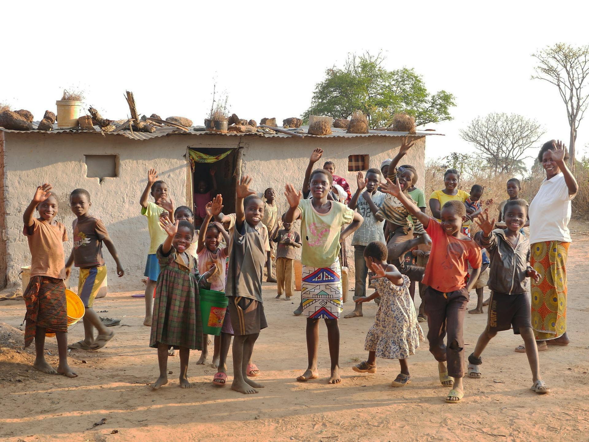 En stor grupp med leende och glada barn med händerna i luften står framför ett litet hus.