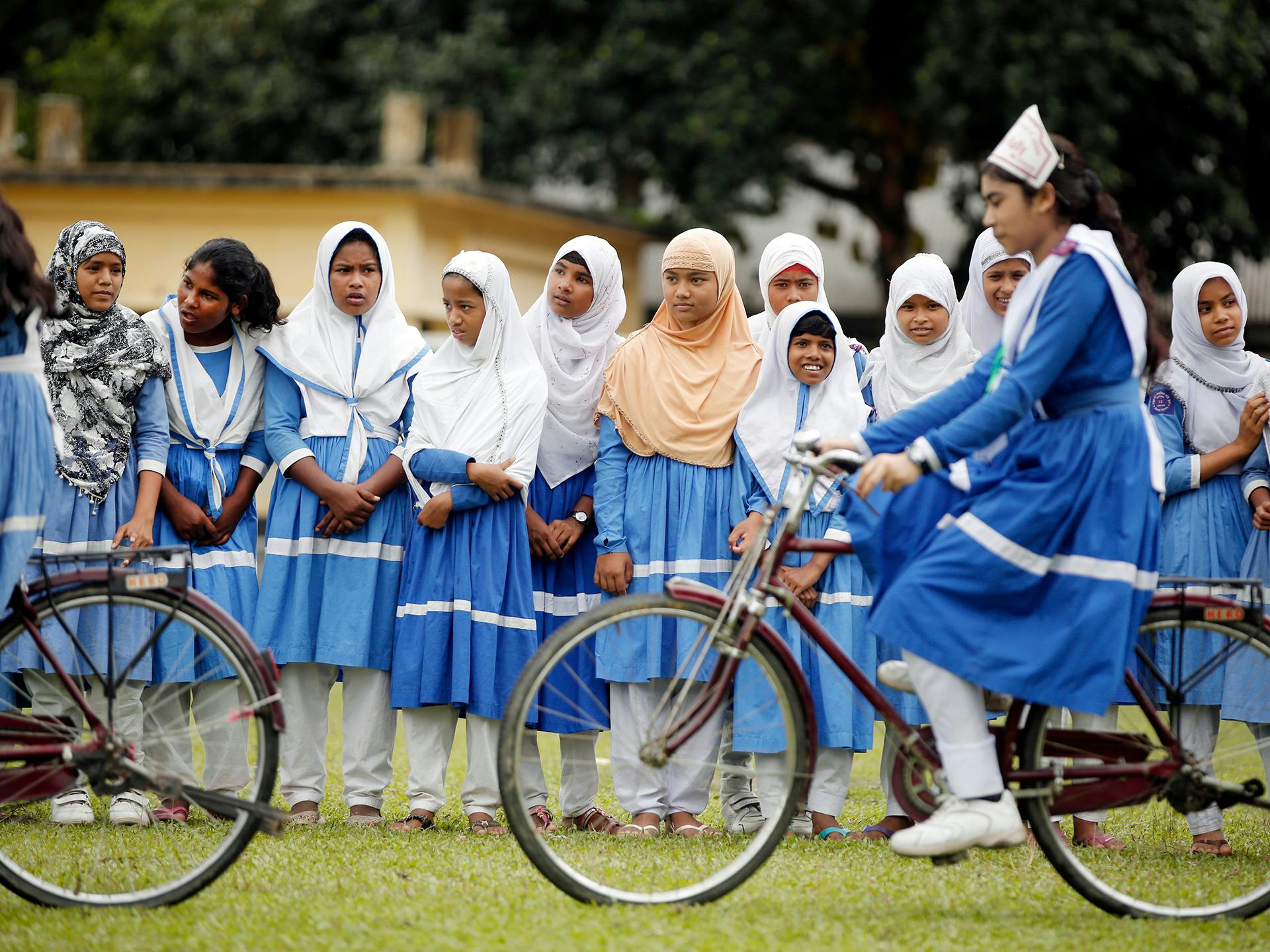 En grupp av flickor och unga kvinnor i likadana skoluniformer ser på när två unga kvinnor cyklar framför dom.