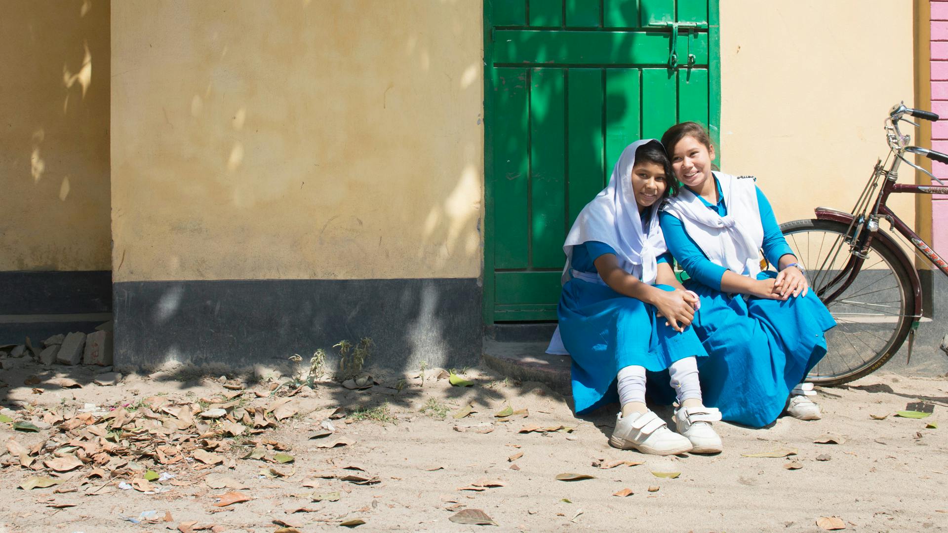 Två flickor i skoluniform sitter framför ett hus. Det står en cykel bredvid.