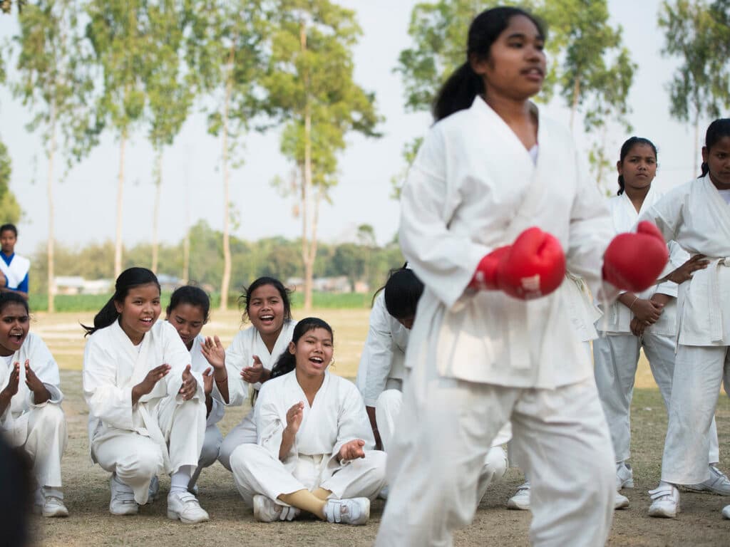 En grupp unga kvinnor sitter på marken och hejar på en annan kvinna som tränar karate.