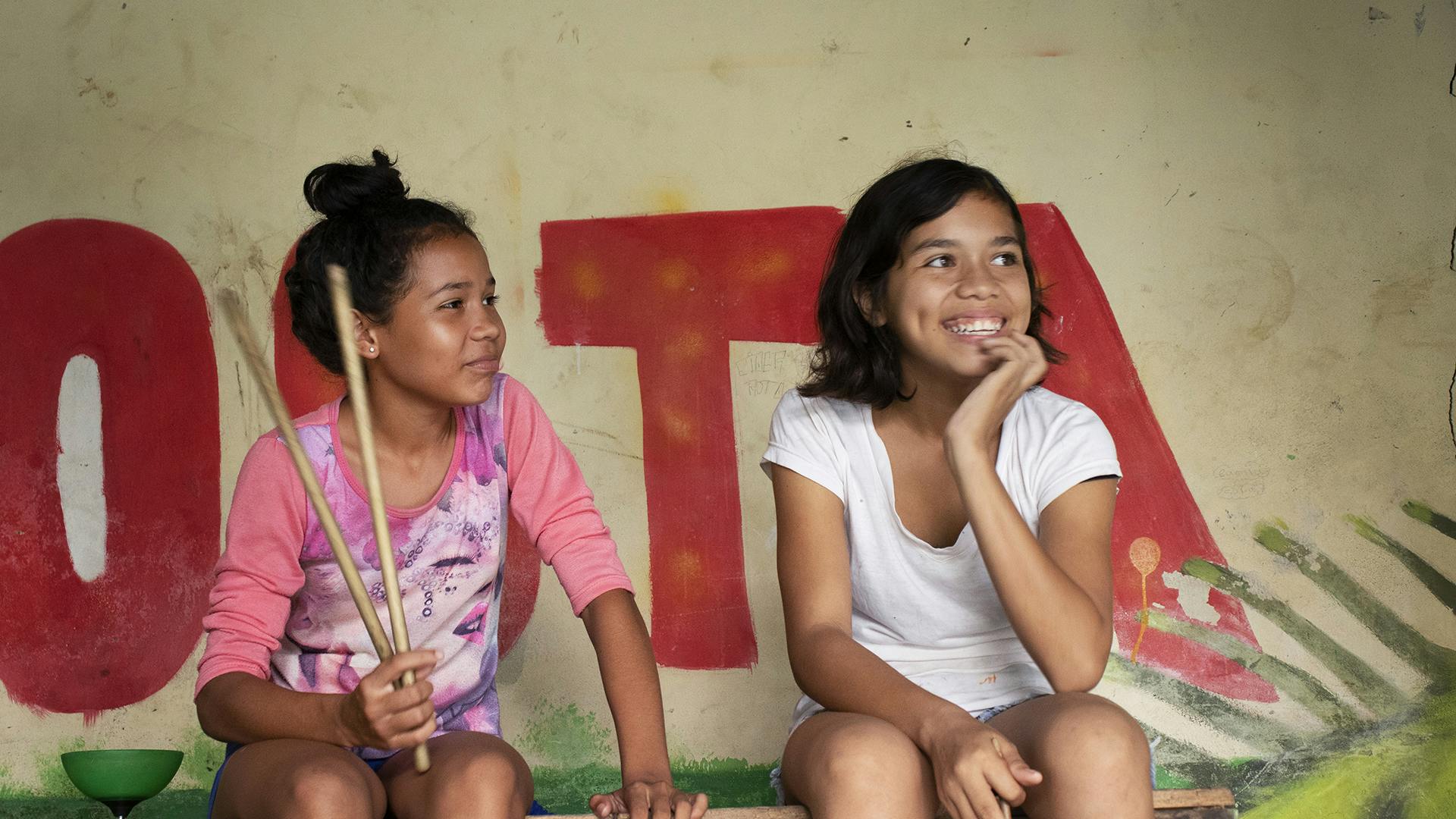 Två flickor sitter och skrattar, en av dom har två stavar i handen.