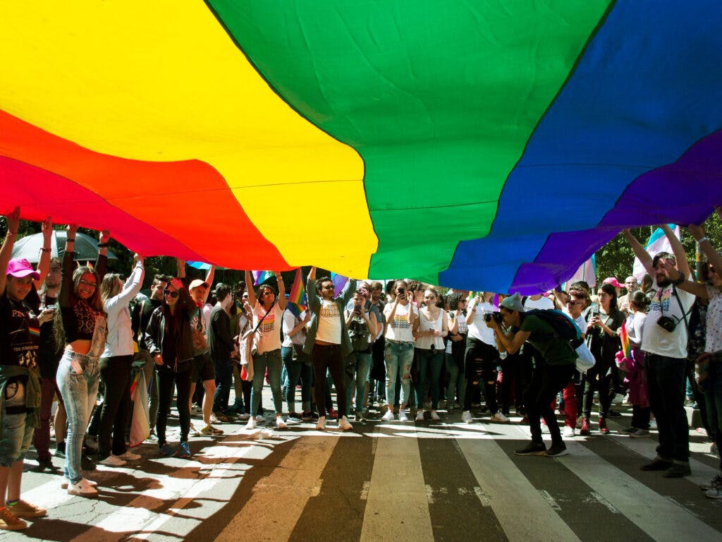 En stor regnbågsflagga täcker halva bilden. Under flaggan syns många människor som håller flaggan med sina händer.