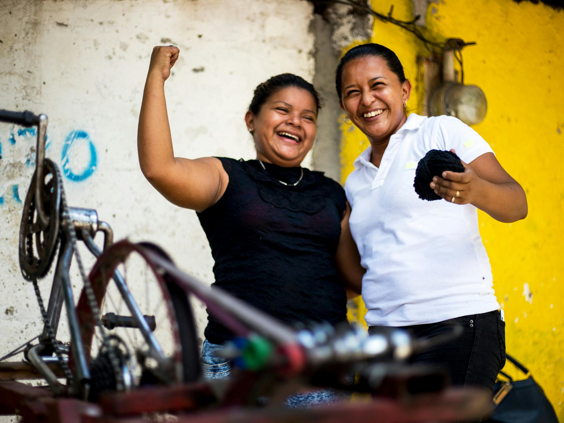 Två skrattande kvinnor står framför en uppochner-vänd cykel. Den ena kvinnan höjer armen med knuten näve.