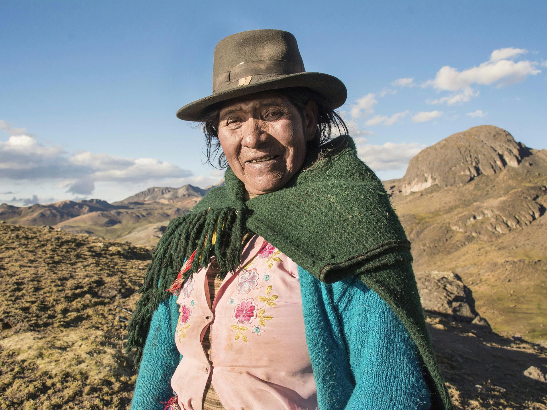 En äldre kvinna med hatt och sjal om axlarna står i ett landskap med berg och torra slätter.