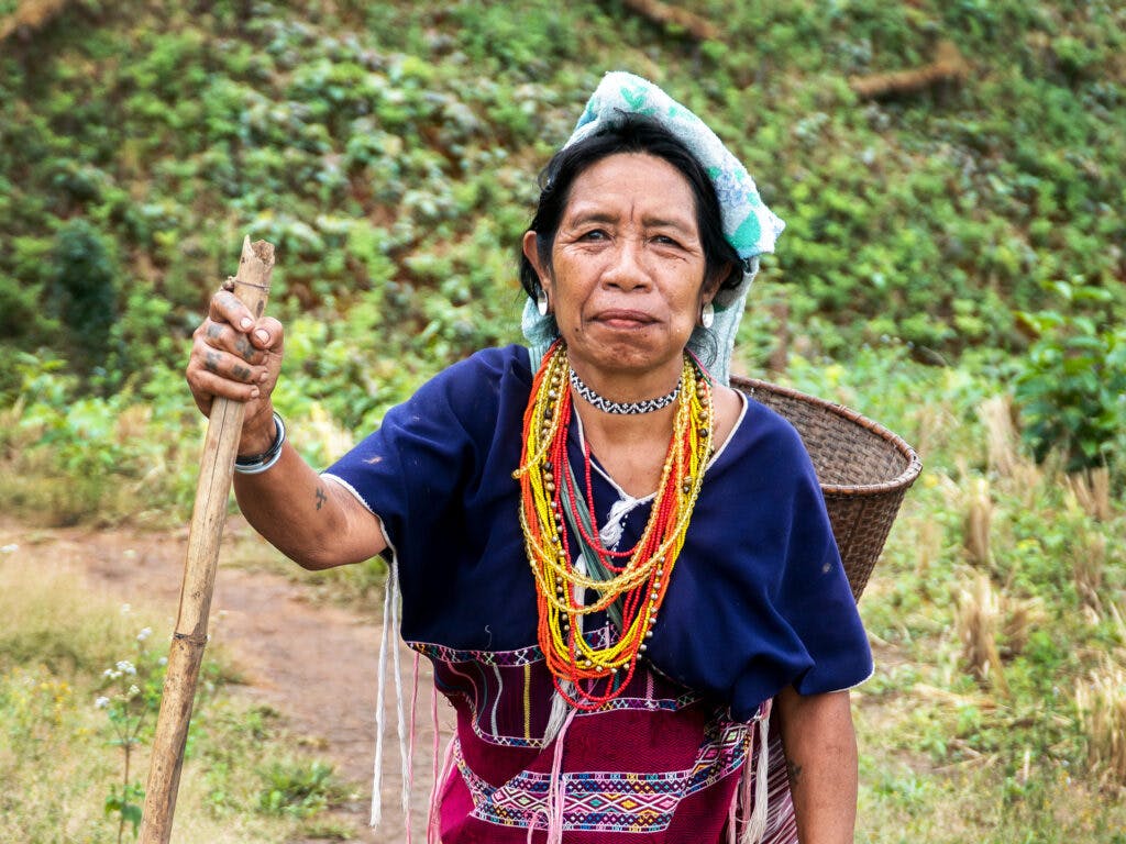 En thailändsk äldre kvinna med färgglada smycken står i ett grönt fält med en stav i handen. På ryggen har hon en korg.
